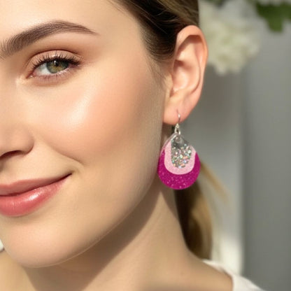 Close-up of a woman wearing Pink Glow resin dangle earrings in small size pink and purple design against a blurred background.