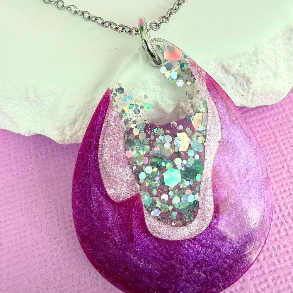 Close up of handmade resin necklace with a shimmery purple and silver pendant on a silver toned chain resting against a white stone on a purple background.
