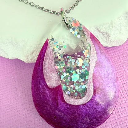 Close up of handmade resin necklace with a shimmery purple and silver pendant on a silver toned chain resting against a white stone on a purple background.