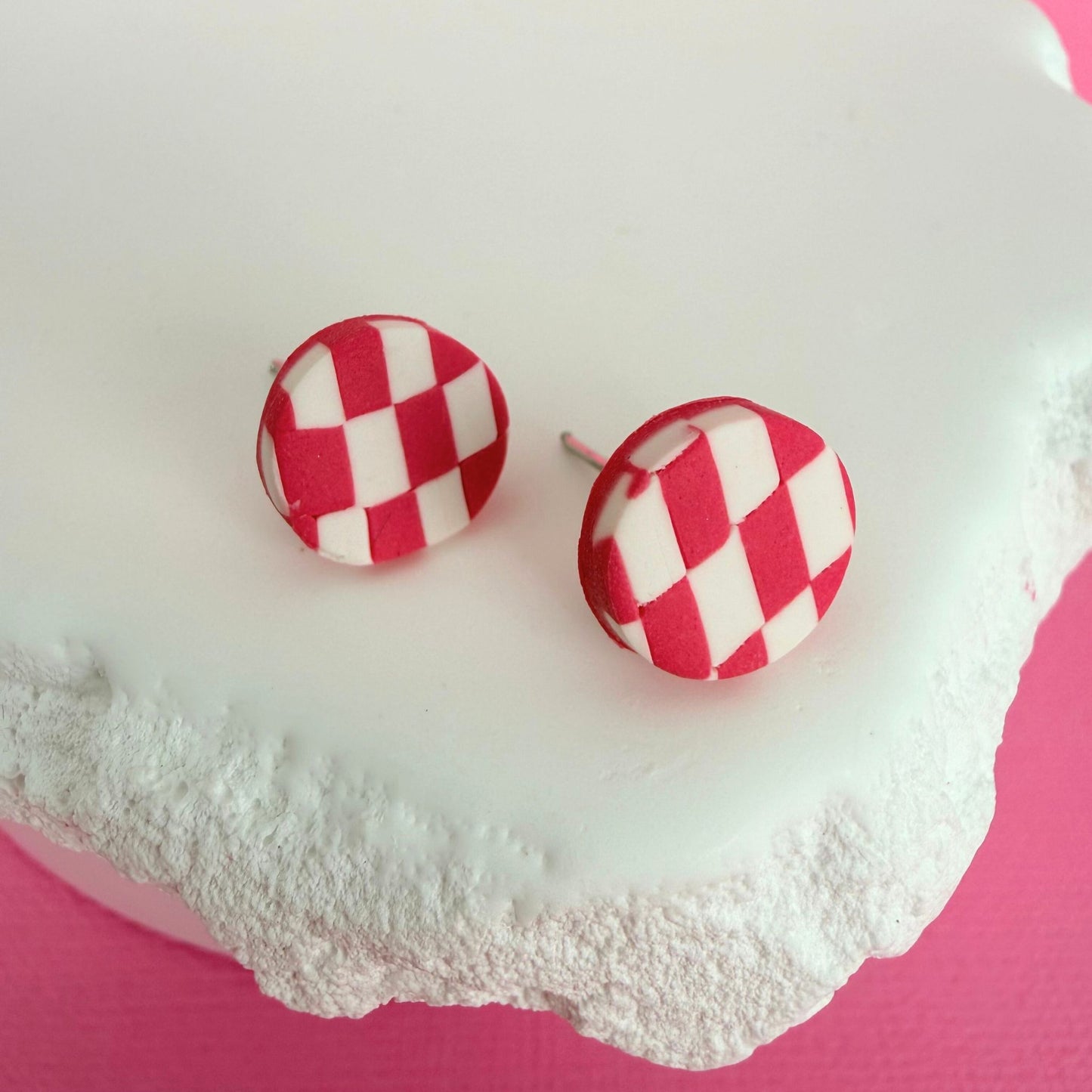 Pink and white checkered polymer clay stud earrings on a textured white surface with a pink background.