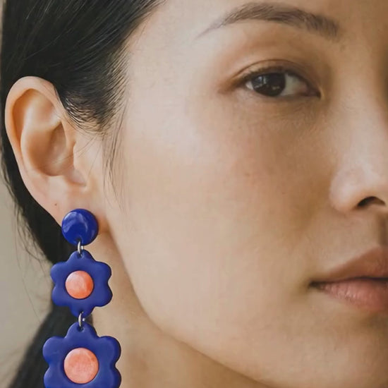 Woman wearing a blue top and coral pleated skirt gently moving, with camera zooming in to highlight bold statement coral and blue flower earrings adding personality to an everyday outfit.