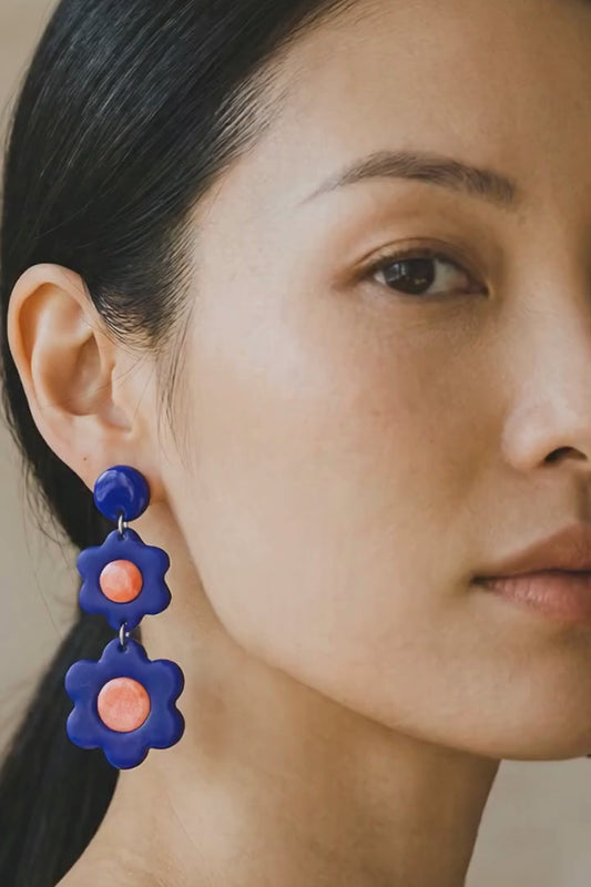 Woman wearing a blue top and coral pleated skirt gently moving, with camera zooming in to highlight bold statement coral and blue flower earrings adding personality to an everyday outfit.