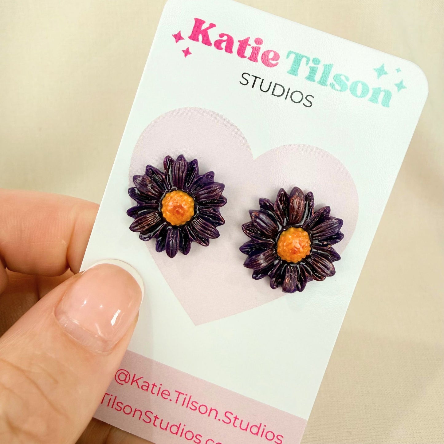 Purple daisy-shaped handmade stud earrings on a Katie Tilson Studios earring card held by a hand.