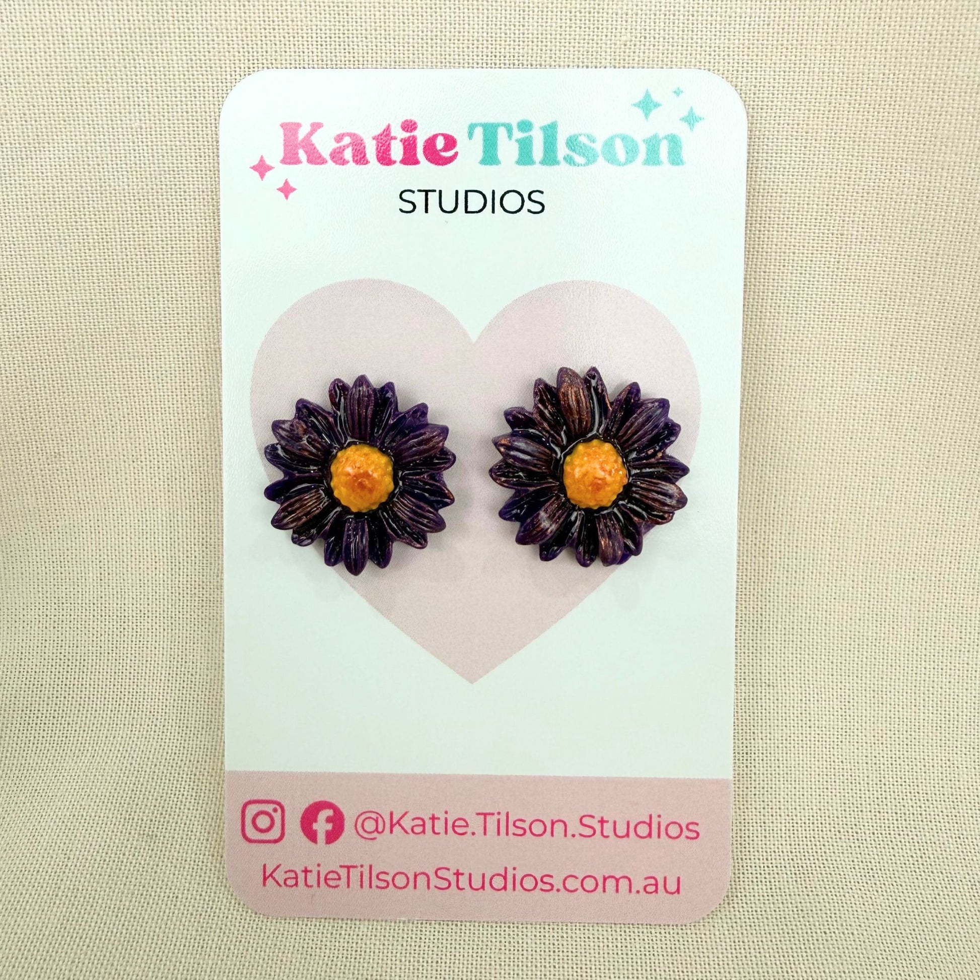 Purple daisy-shaped handmade stud earrings on a Katie Tilson Studios earring card against a beige background.