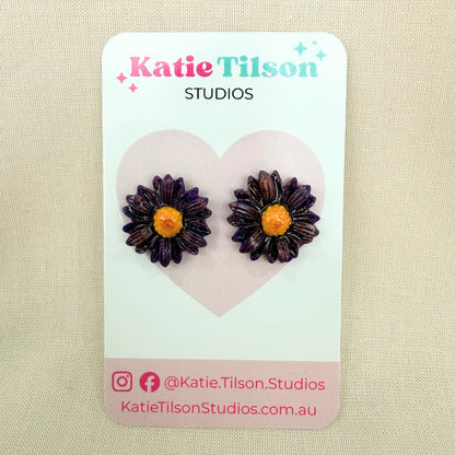 Purple daisy-shaped handmade stud earrings on a Katie Tilson Studios earring card against a beige background.
