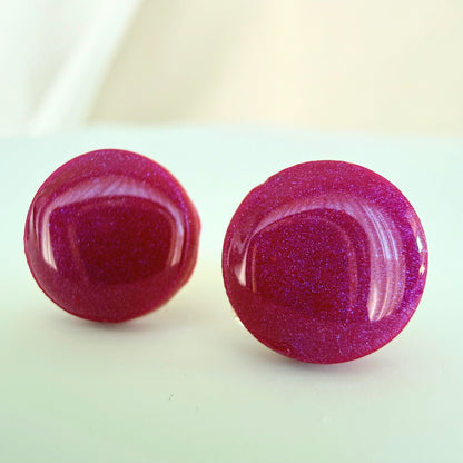 A pair of rich raspberry coloured circle studs with shimmer and glossy resin finish resting on a white surface.