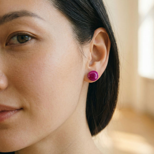 Close-up of a woman wearing round raspberry tone stud earrings with a glossy finish against a blurred background.