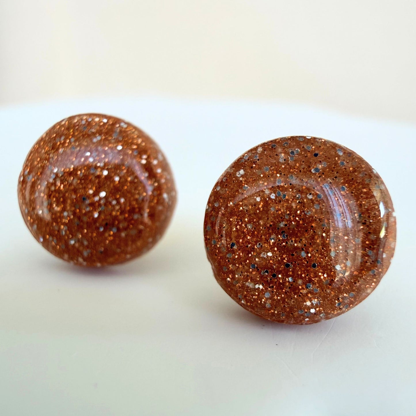 A pair of warm rose gold colour circle studs with silver and gold shimmer and glossy resin finish resting on a white surface.