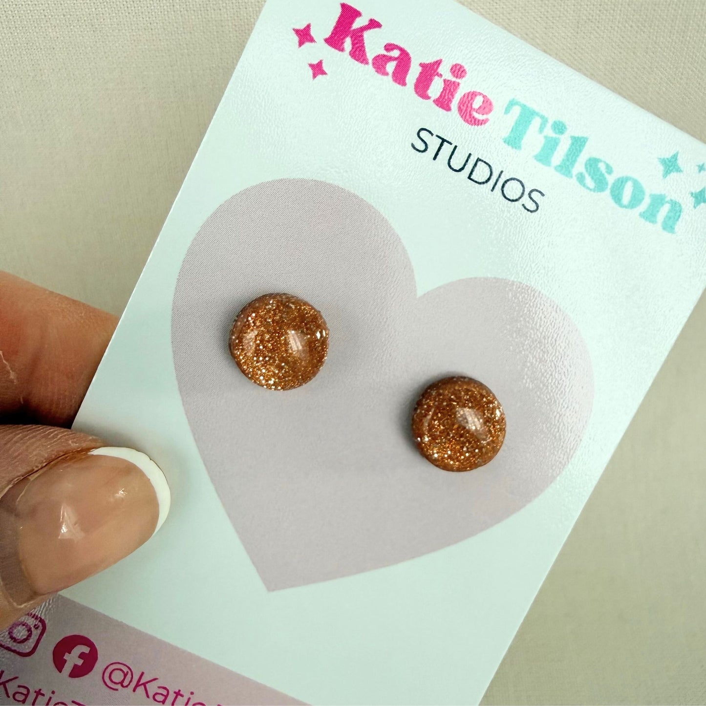 Rose gold glitter circular stud earrings on a Katie Tilson Studios card held by a hand.