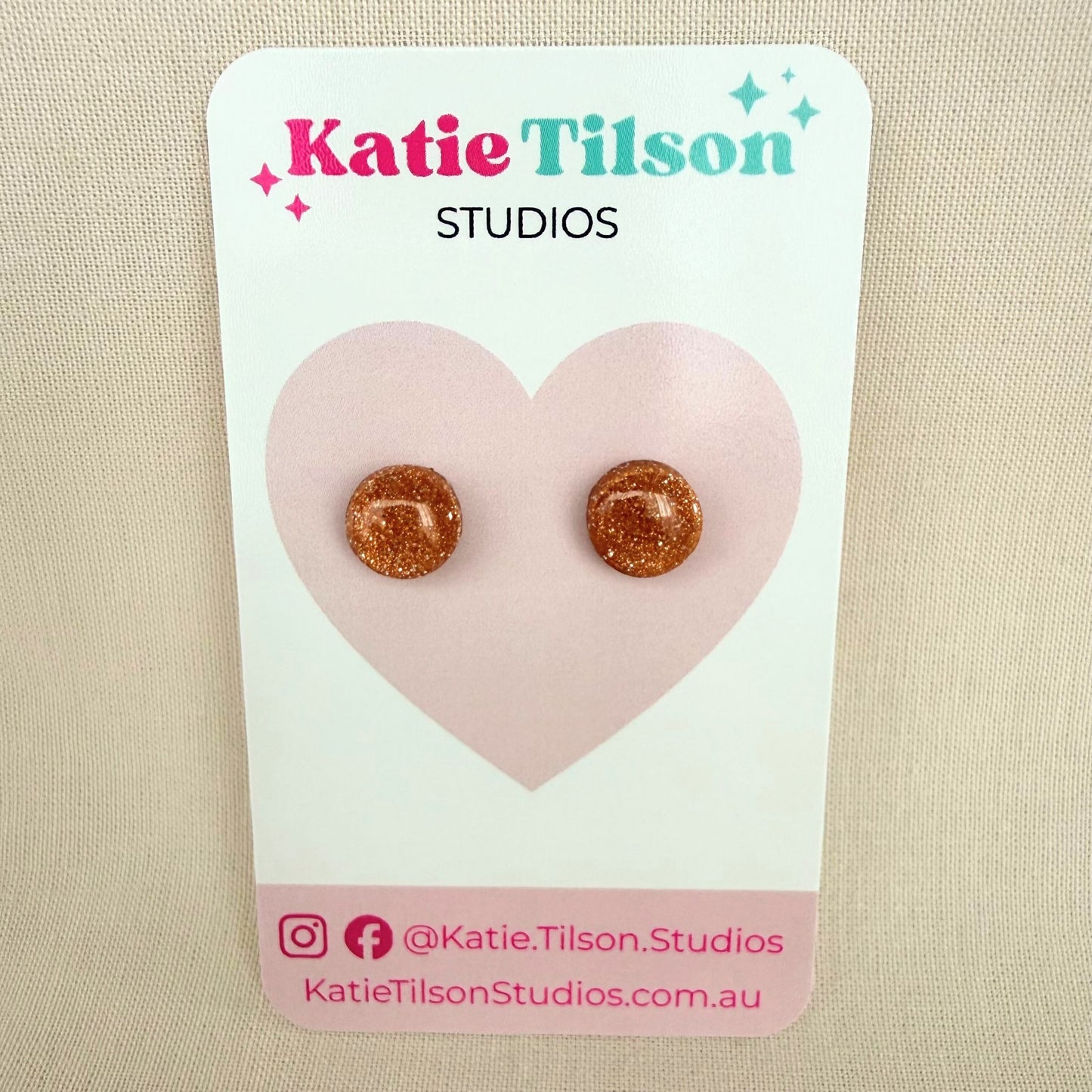 Rose gold glitter circular stud earrings on a Katie Tilson Studios card against a beige background.