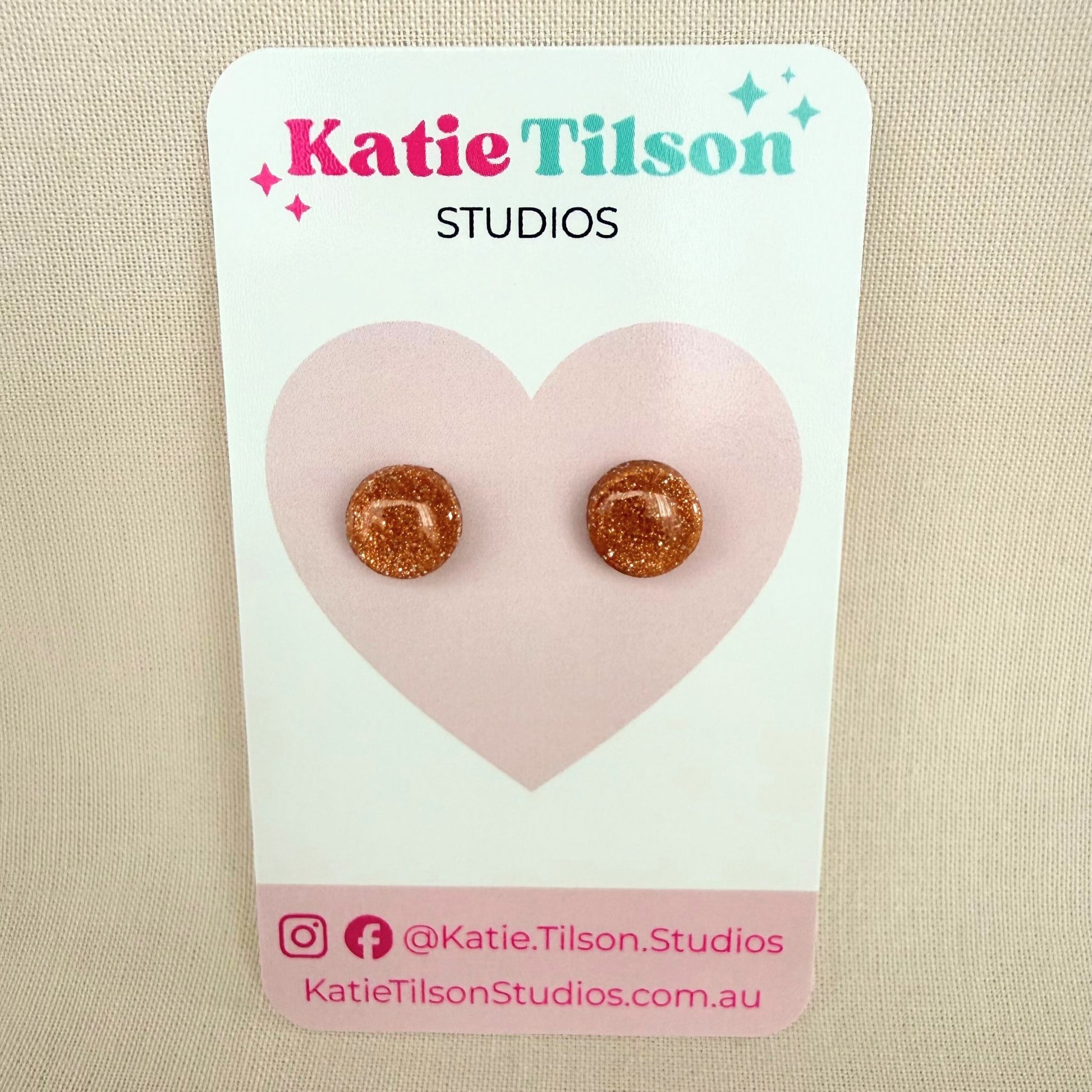 Rose gold glitter circular stud earrings on a Katie Tilson Studios card against a beige background.