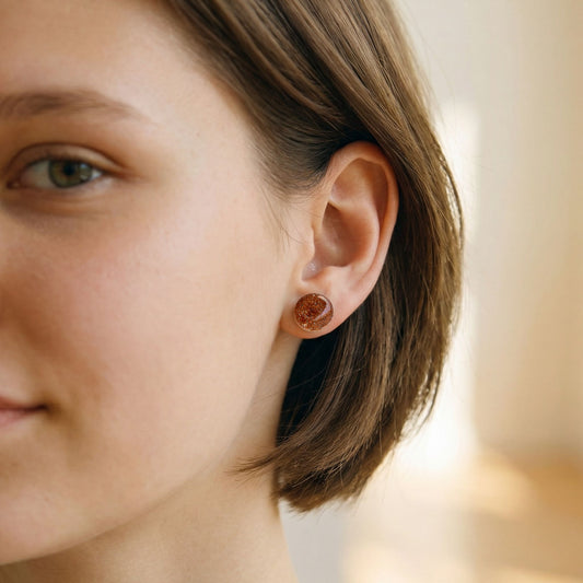 Close-up of a woman wearing round rose gold tone stud earrings with a glossy finish against a blurred background.
