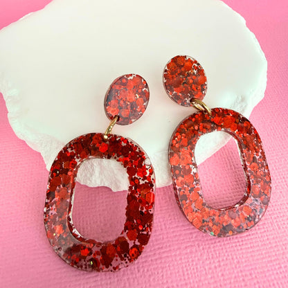 Australian handmade resin drop earrings in red glitter design resting against a white stone with a pink background.