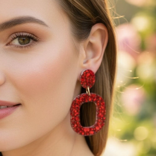 Close-up of a woman wearing Ruby Glow Australian handmade resin drop earrings in a red glitter design with a blurred outdoor background.