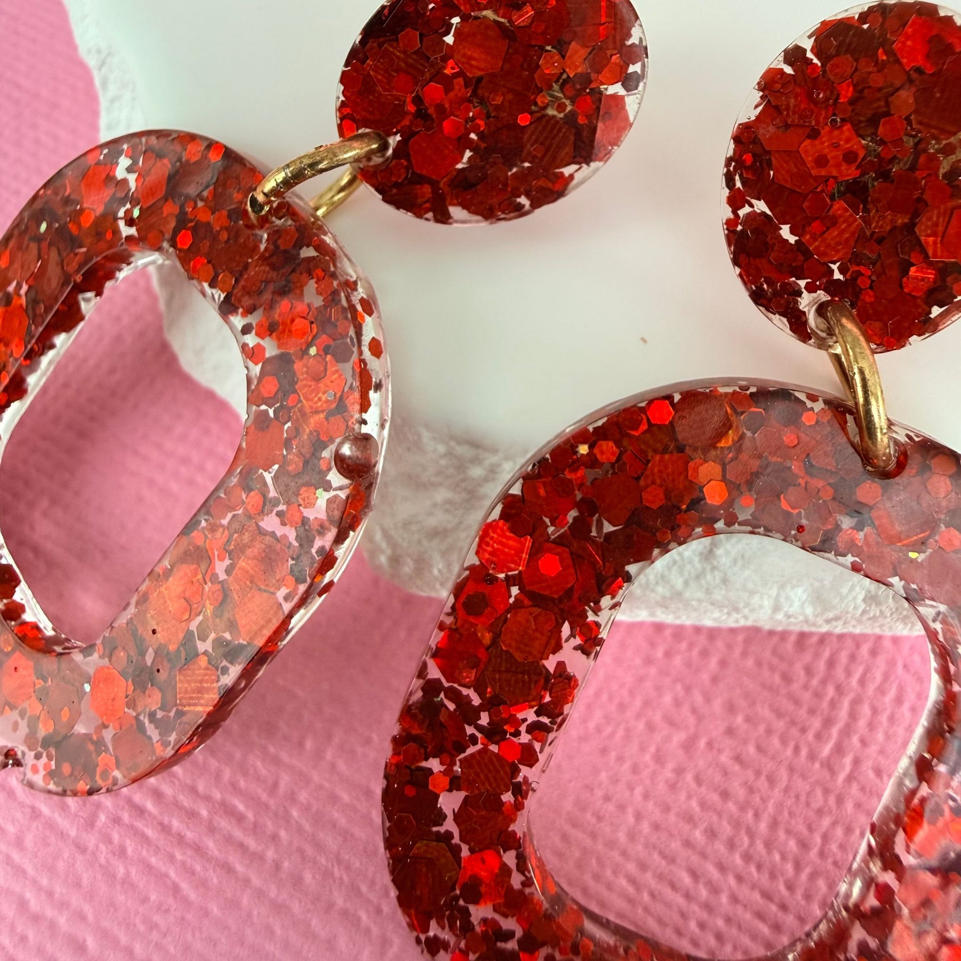 Close up of Ruby Glow Australian handmade resin drop earrings in red glitter design resting against a white stone with a pink background.