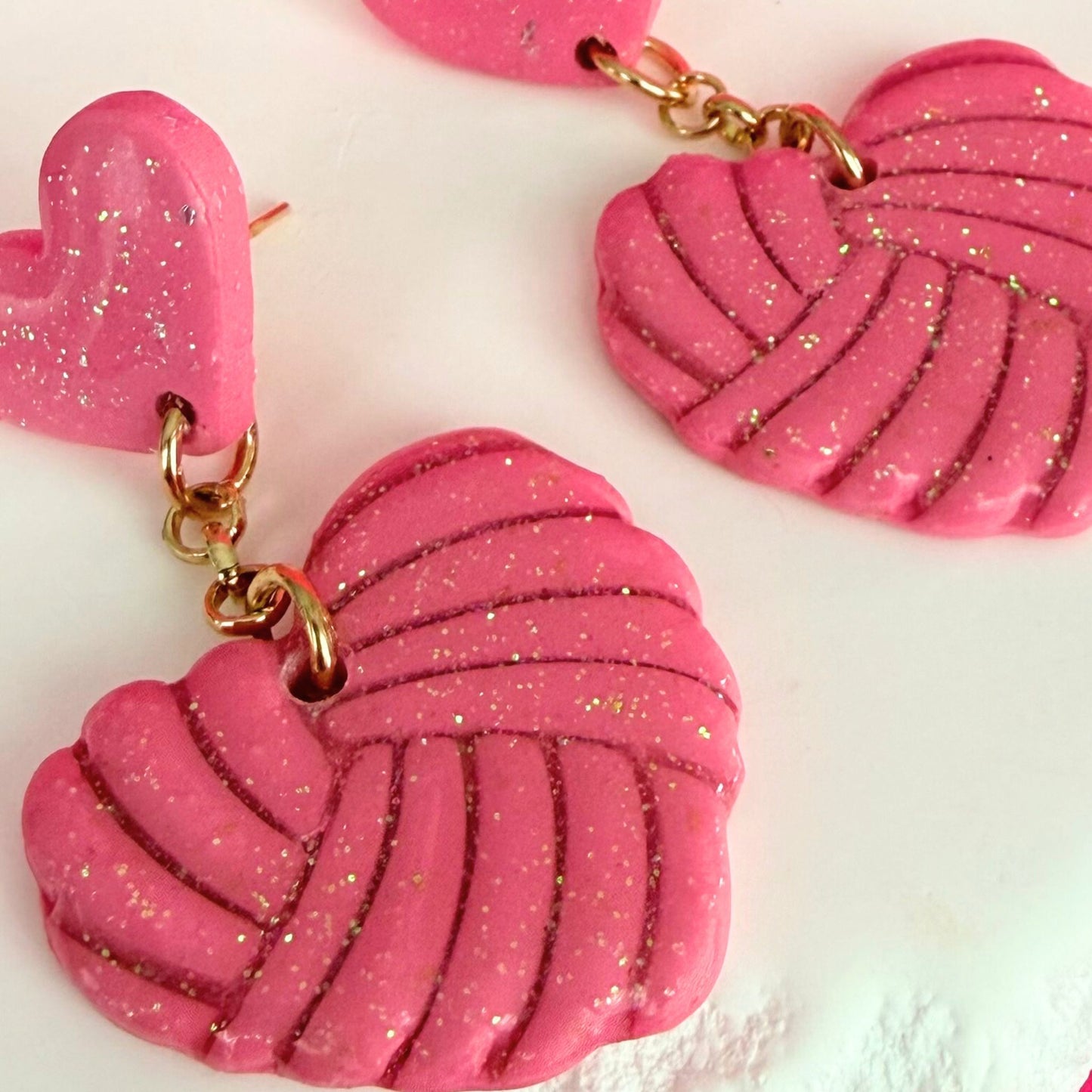 Pink heart-shaped drop earrings with knotted texture and gold accents on a white background.