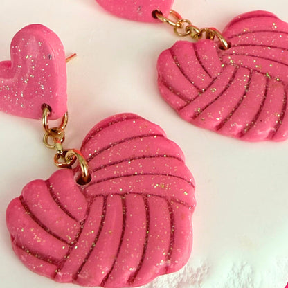 Pink heart-shaped drop earrings with knotted texture and gold accents on a white background.