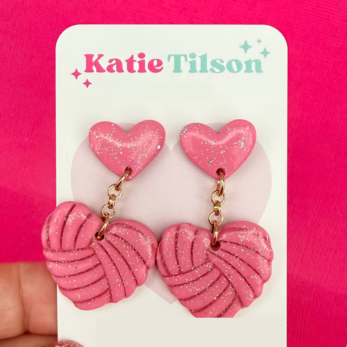 Pink heart-shaped drop earrings with a knitted heart shaped texture on a Katie Tilson branded card against a pink background.