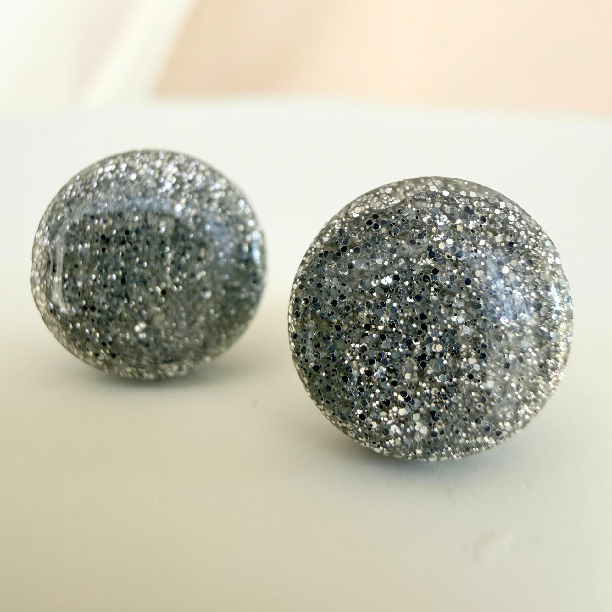 Glossy silver stud earrings with silver glitter in a circular shape design resting on a white surface.
