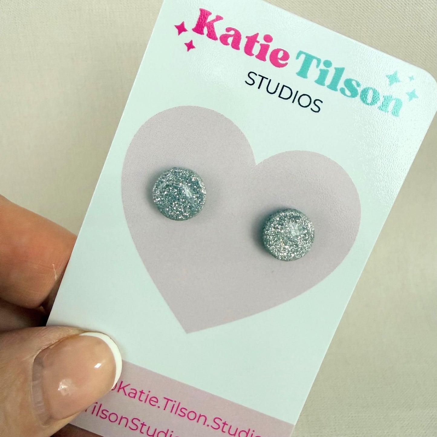 Pair of silver glittery circular stud earrings on a Katie Tilson Studios card held by a hand.
