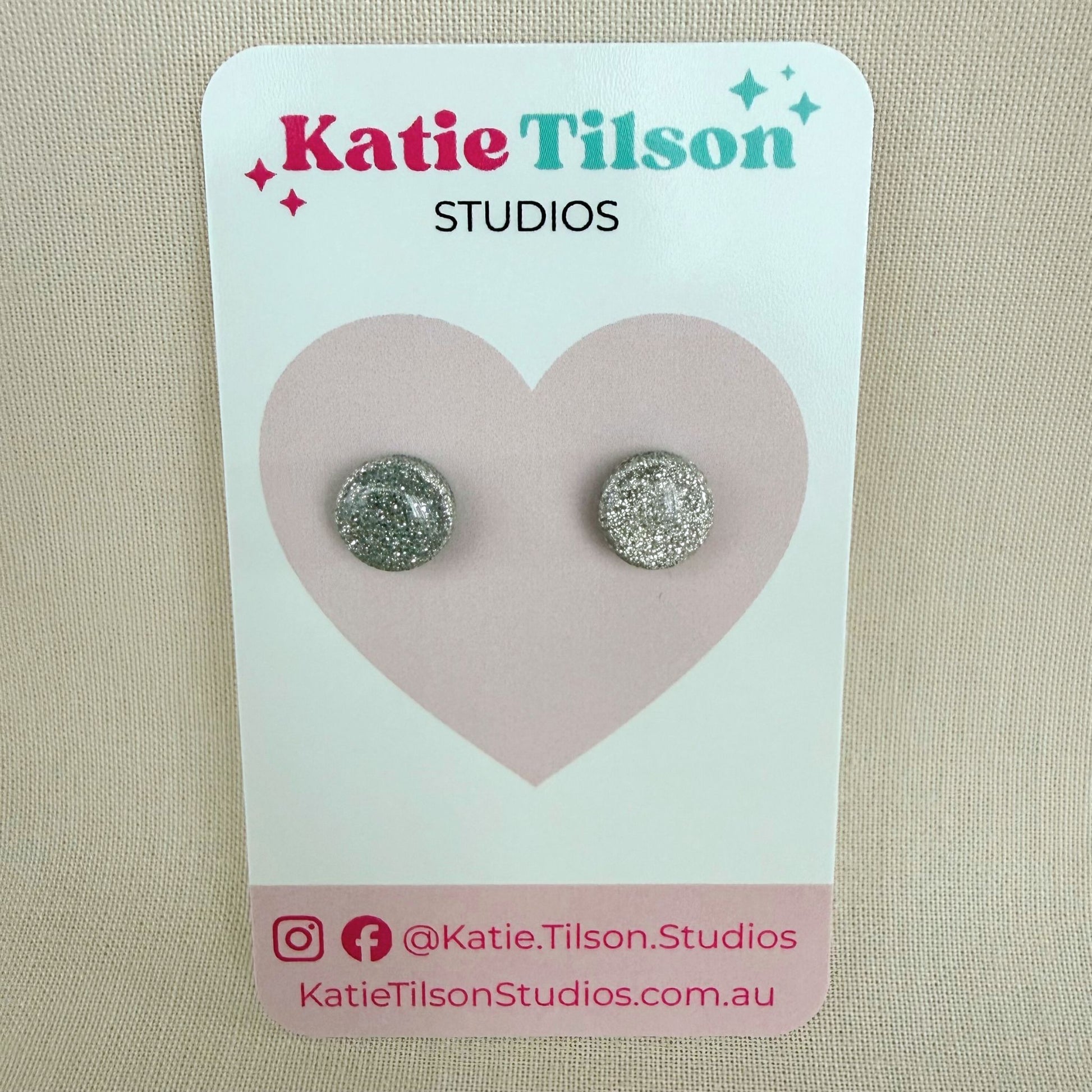 Pair of silver glittery circular stud earrings on a Katie Tilson Studios card against a beige background.