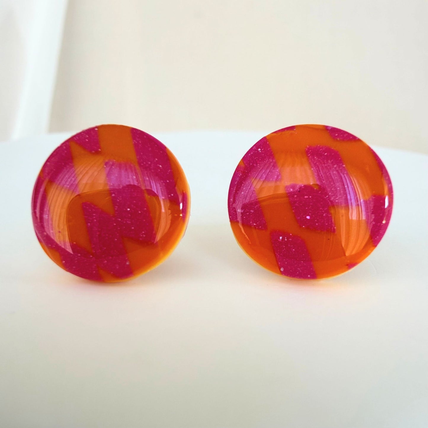 A pair of bright orange and pink checkered pattern circle studs with shimmer and glossy resin finish resting on a white surface.