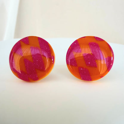 A pair of bright orange and pink checkered pattern circle studs with shimmer and glossy resin finish resting on a white surface.