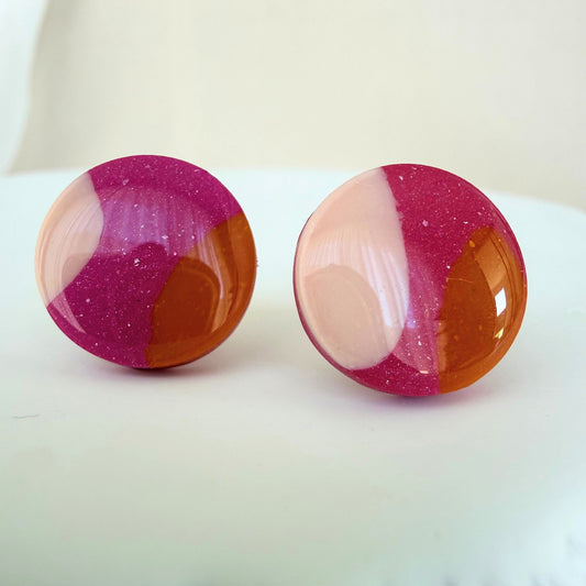 A pair of pink, orange and bright pink abstract design circle studs with shimmer and a glossy resin finish resting on a white surface.