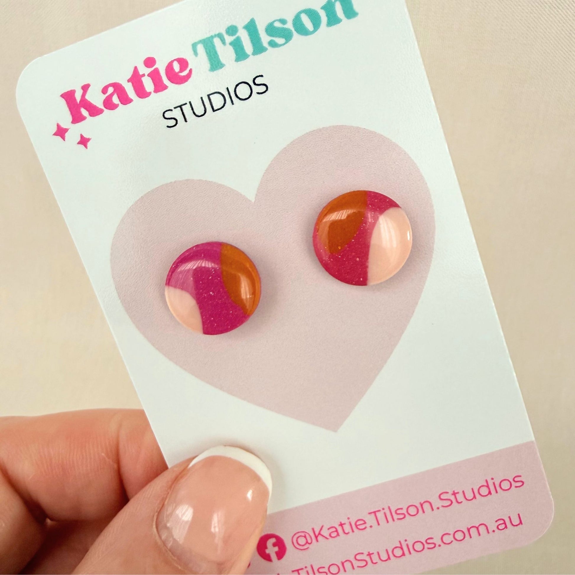 Pair of dark pink, orange and pastel pink colorful stud earrings on a Katie Tilson Studios card held by a hand.