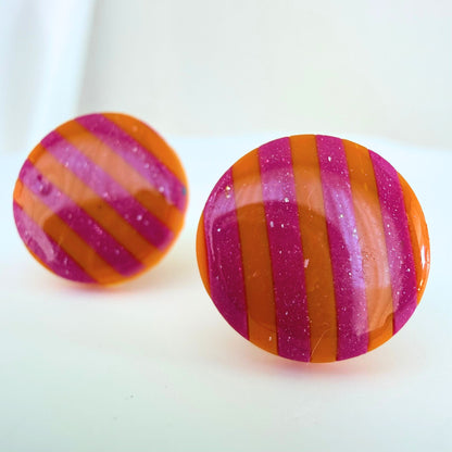 A pair of bright orange and pink striped circle studs with shimmer and glossy resin finish resting on a white surface.
