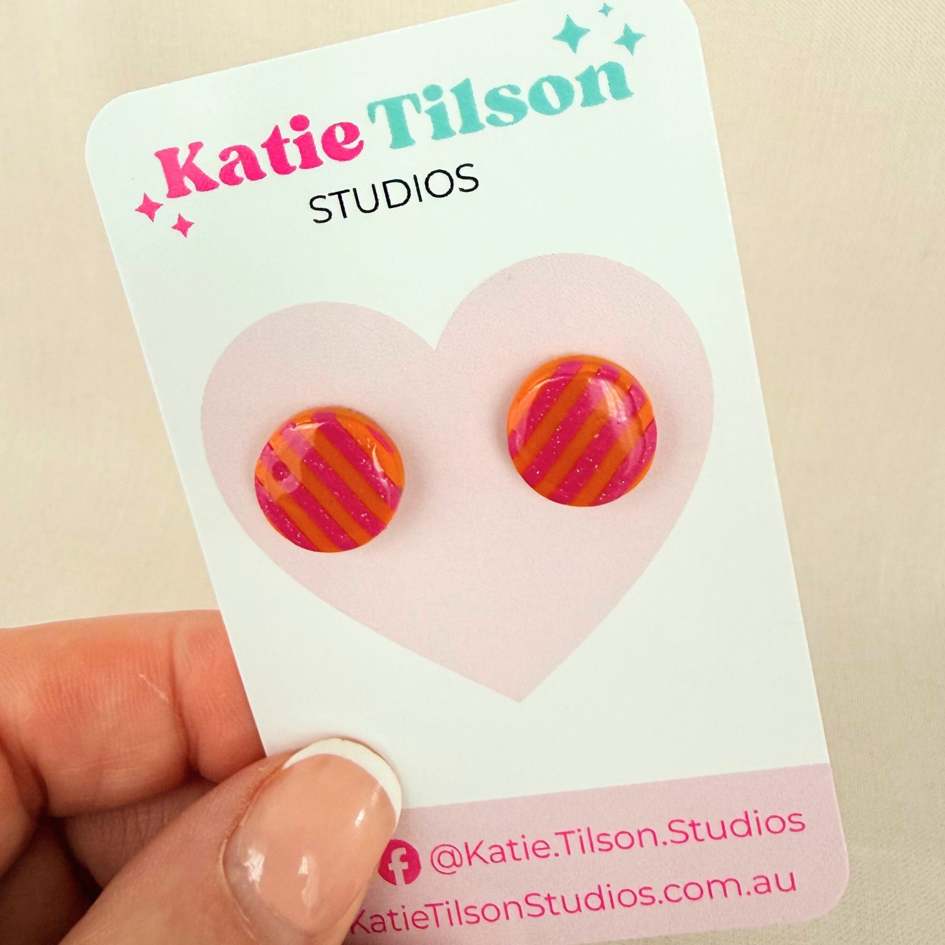 Colorful pink and orange striped circular stud earrings on a Katie Tilson Studios card held by a hand.