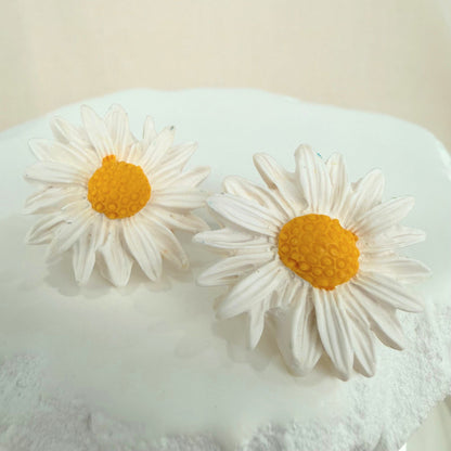 White daisy-shaped handmade stud earrings with yellow centers on a white stone surface against a beige background.