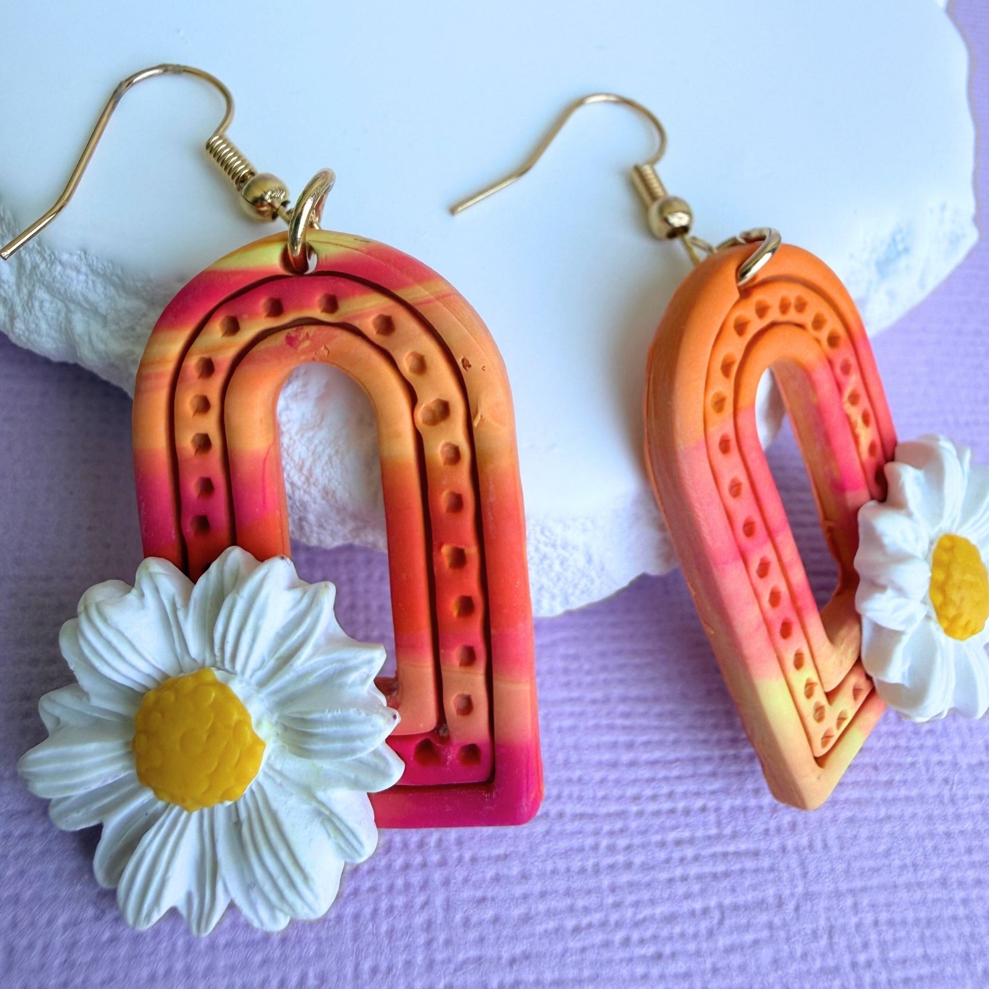 Colorful arch-shaped handmade dangle earrings with daisy flowers on a white and purple background.