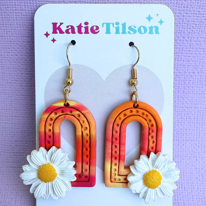 Colorful arch-shaped earrings with daisy flowers on a Katie Tilson branded card against a purple background.