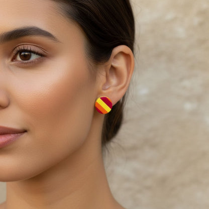 Close-up of a woman wearing colorful Sunset Pop striped stud earrings in red, orange and yellow against a neutral background.