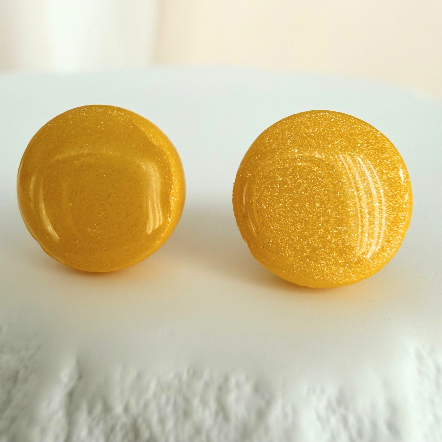 A pair of bright sunshine yellow circle studs with shimmer and glossy resin finish resting on a white surface.