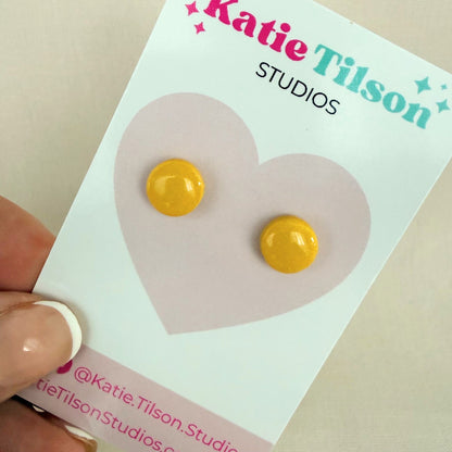 Sunshine yellow circular stud earrings on a Katie Tilson Studios earring card held by a hand.