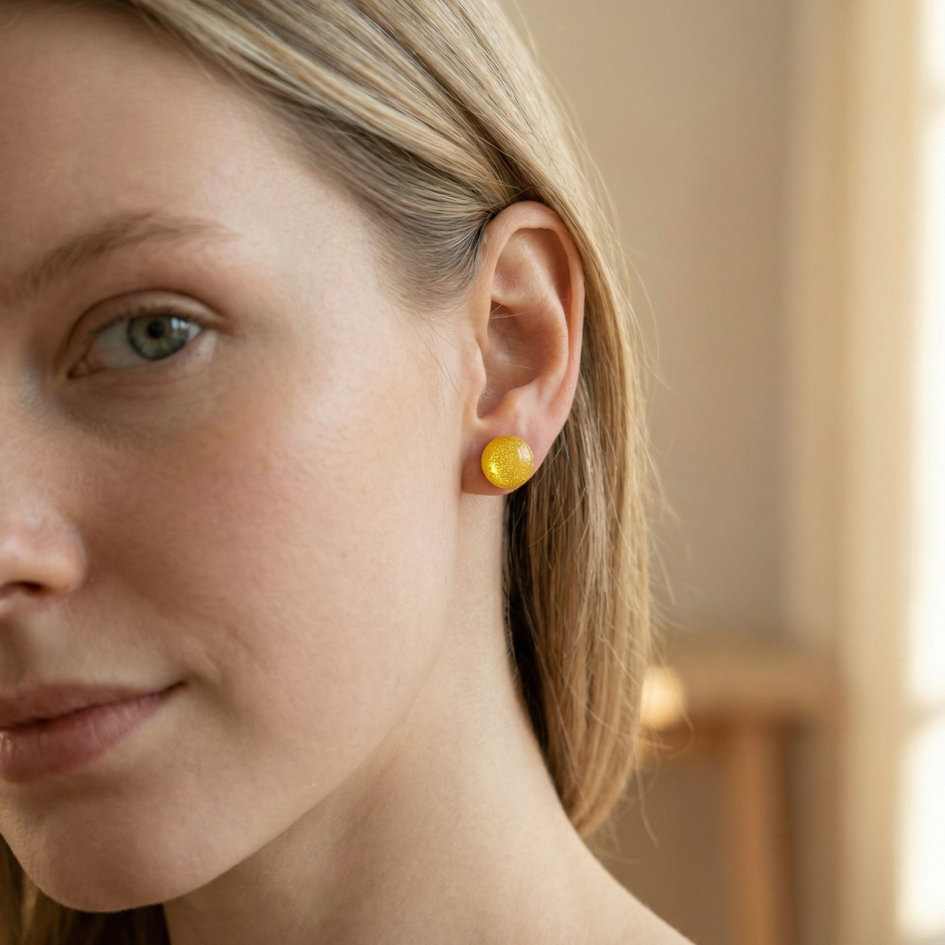 Close-up of a woman wearing round sunshine yellow stud earrings with a glossy finish against a blurred background.