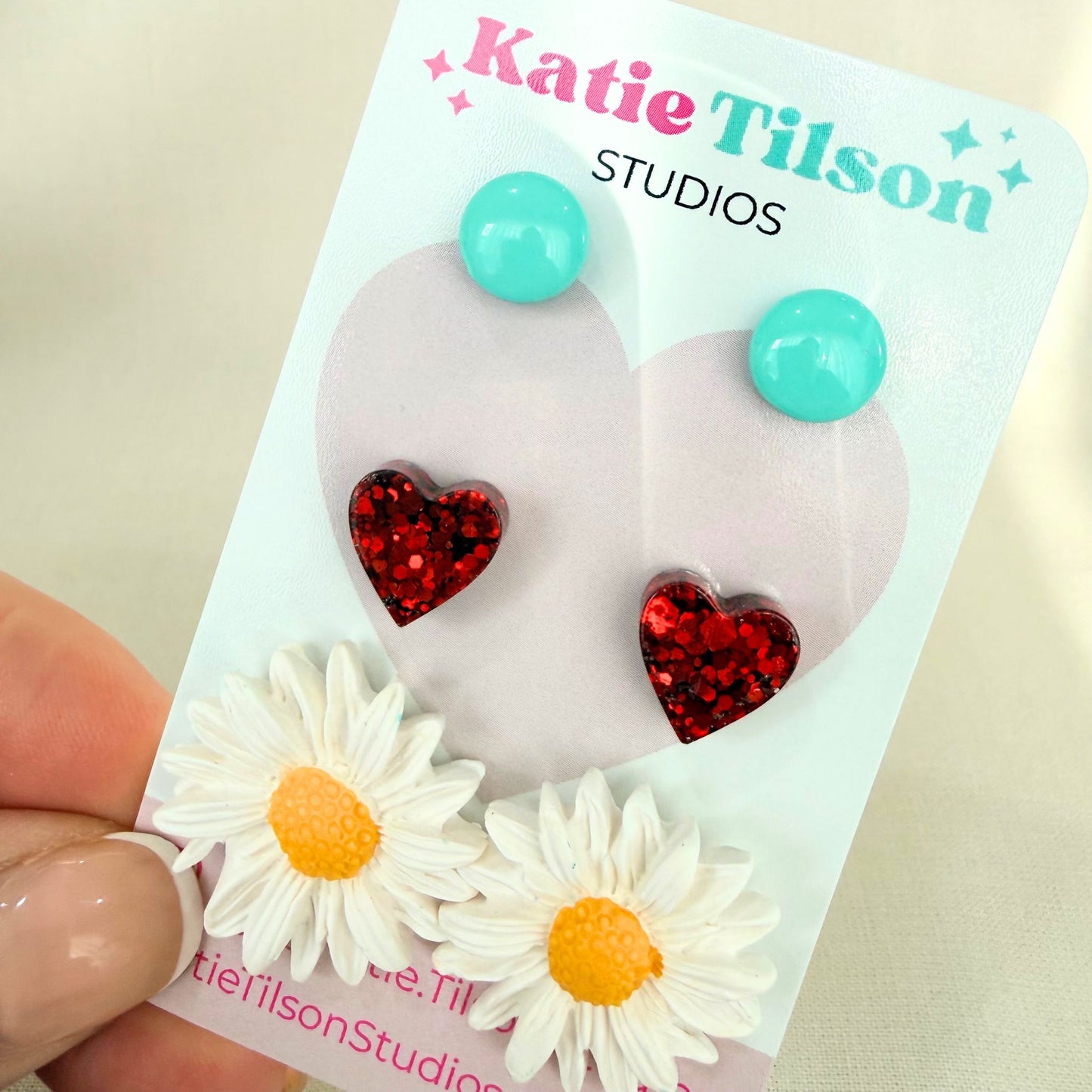 Mint circular studs, red heart studs and white daisy studs displayed on a Katie Tilson Studios card held by a hand.