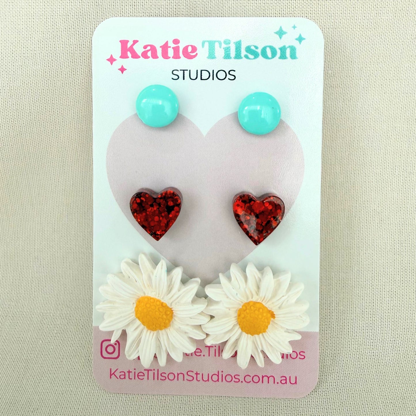 Mint circular studs, red heart studs and white daisy studs displayed on a Katie Tilson Studios card against a beige background.
