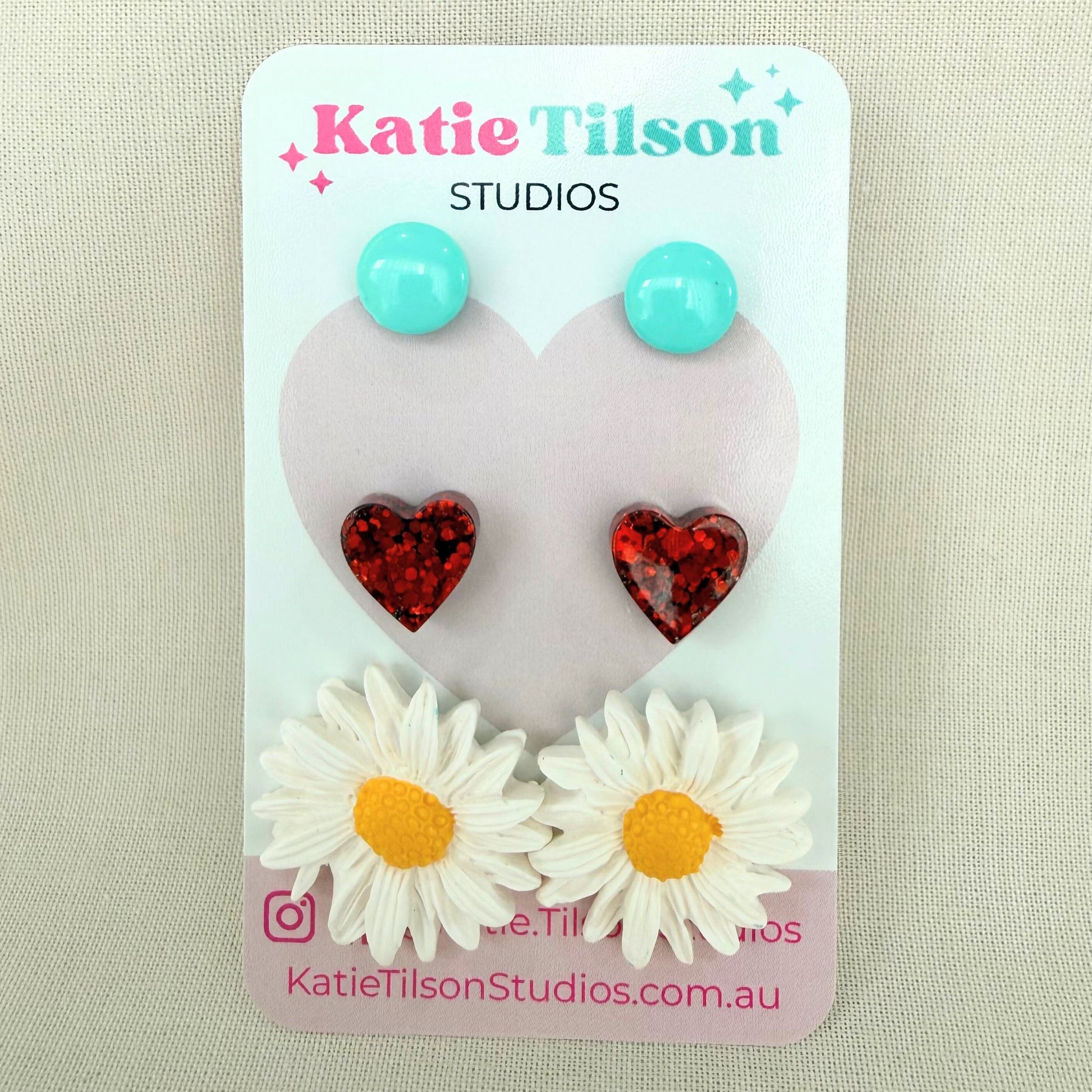 Mint circular studs, red heart studs and white daisy studs displayed on a Katie Tilson Studios card against a beige background.