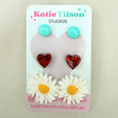 Mint circular studs, red heart studs and white daisy studs displayed on a Katie Tilson Studios card against a beige background.