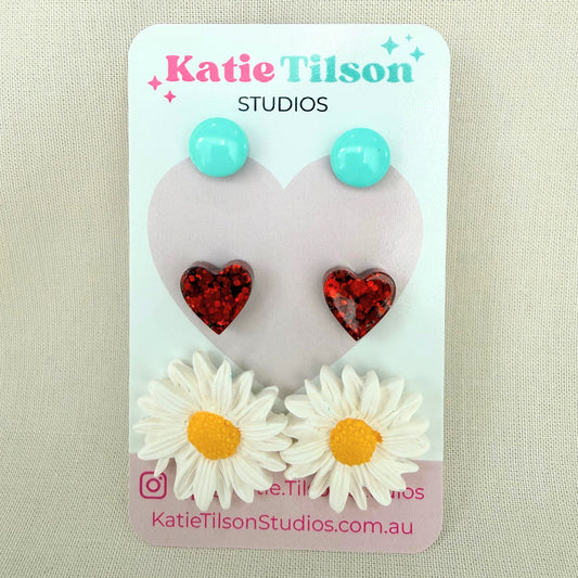 Mint circular studs, red heart studs and white daisy studs displayed on a Katie Tilson Studios card against a beige background.