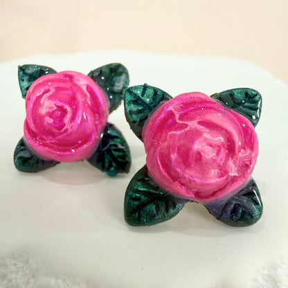 Pair of pink rose stud earrings with green leaves on a white surface.