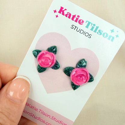 A pair of candy pink rose shaped stud earrings with green leaves on a Katie Tilson Studios card held by a hand.
