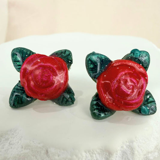 A pair of red rose shaped stud earrings with green leaves on a white surface.