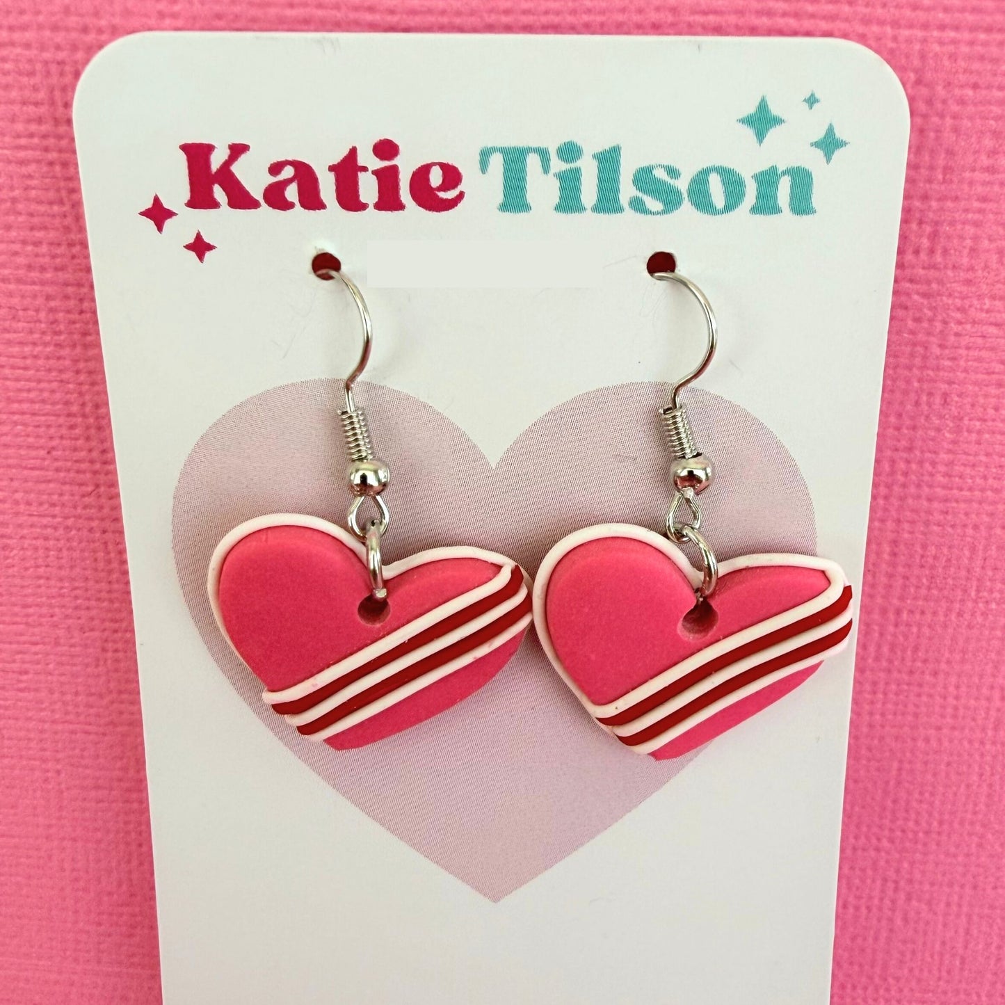 Heart-shaped handmade dangle earrings on a Katie Tilson branded card against a pink background.