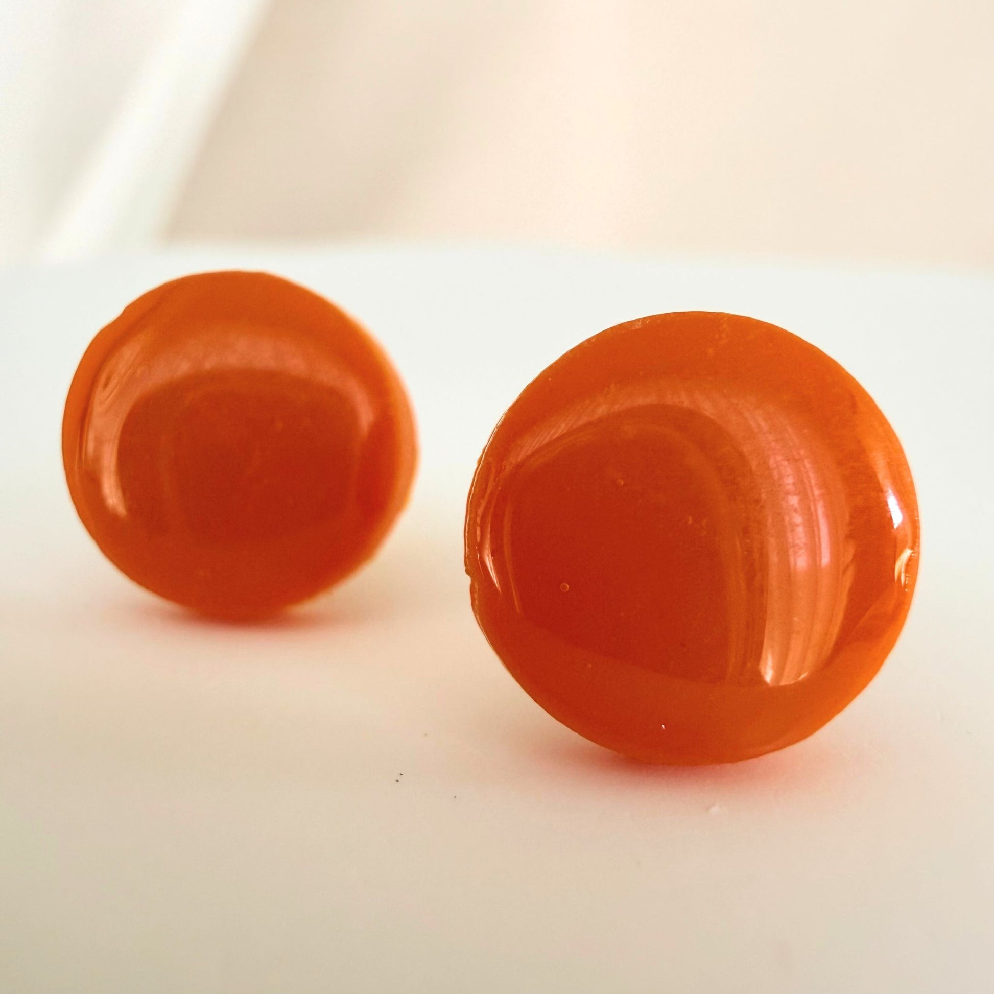 A pair of bright tangerine orange circle studs with a glossy resin finish resting on a white surface.