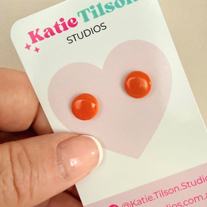 Orange circular stud earrings on a Katie Tilson Studios card held by a hand.