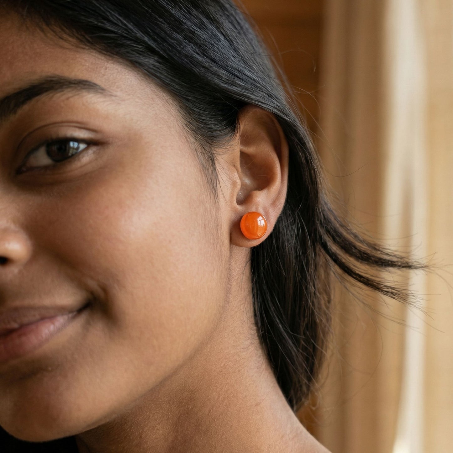 Close-up of a woman wearing round tangerine orange stud earrings with a glossy finish against a blurred background.
